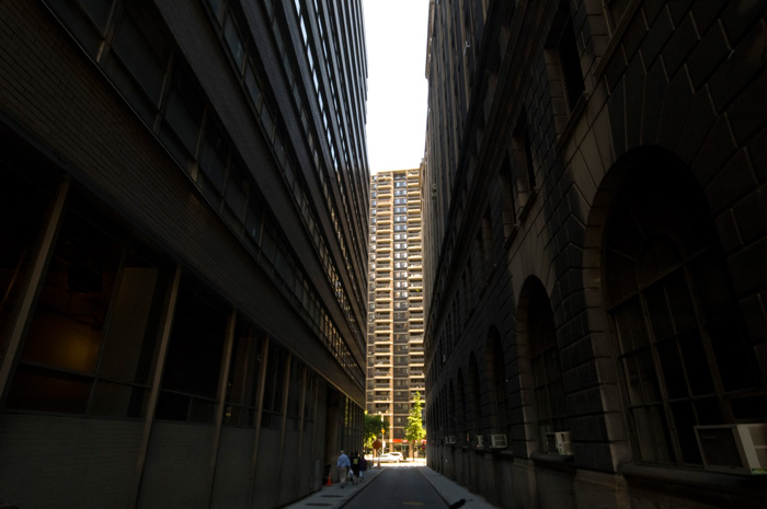 A sliver of light, at the end of the block, fails to shine into the gap between two towering dark buildings.