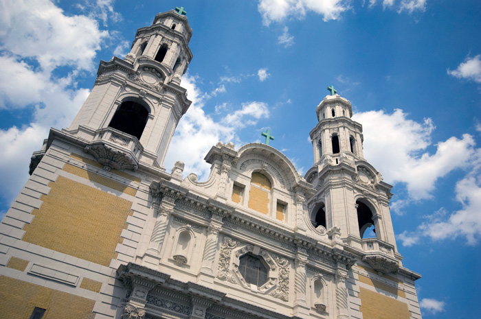 A yellow brick church with white ornamentation, has two tall white steeples at its corners.