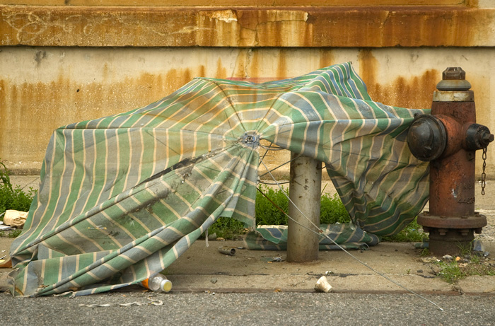 A faded and torn green umbrella lies, destroyed, by a rusting fire hydrant.