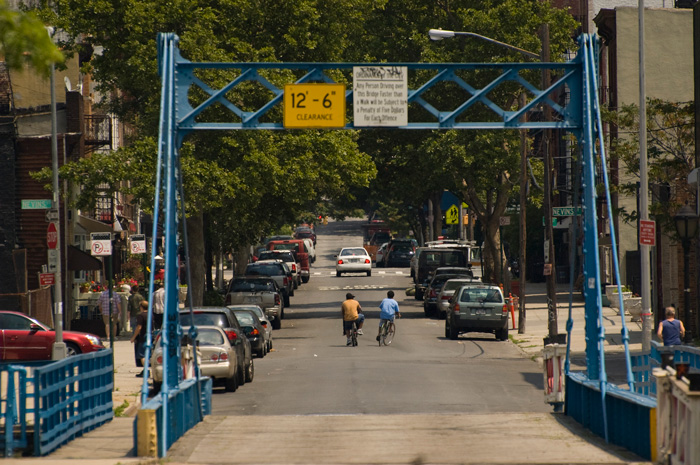 Two teens ride their bikes down the middle of a tree-lined street.