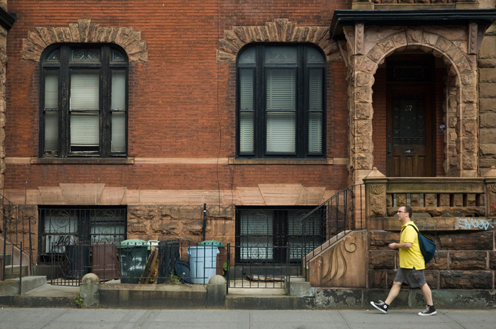 A pedestrian passes in front of a brick home with a stairway and entrance ornamented of stone.