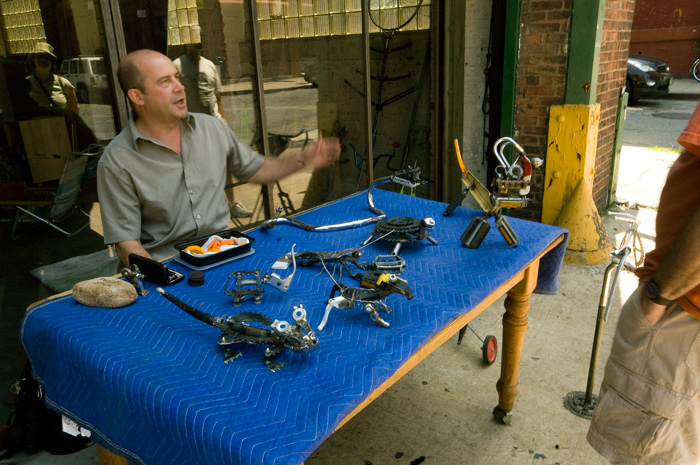 A man sits behind a table displaying several sculptures he's welded out of bicycle parts.