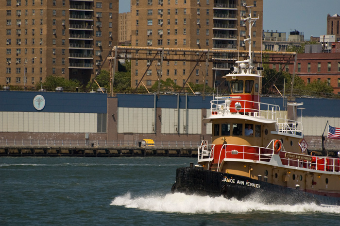 A tan, red, and black tugboat cruising on the river, with tall apartment buildings on the opposite shore.