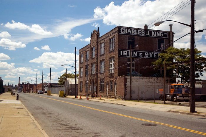 A three story building of brick and stone towers over small industrial buildings nearby, with no one to be seen.