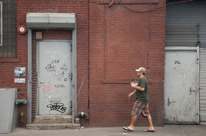 A man in plaid shorts, a t-shirt, and trucker's cap carries coffe cups and a lit cigarette while walking down an industrial-looking street.
