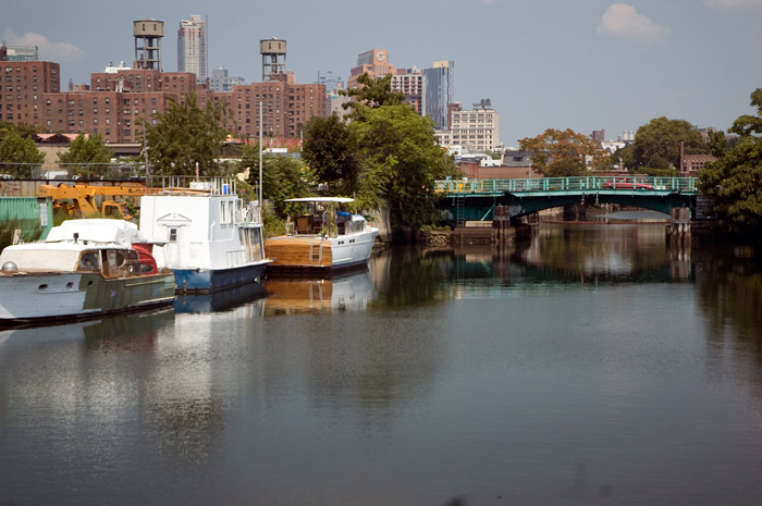Pleasure boats line the side of a canal, and in the background is a bridge and tall buildings.