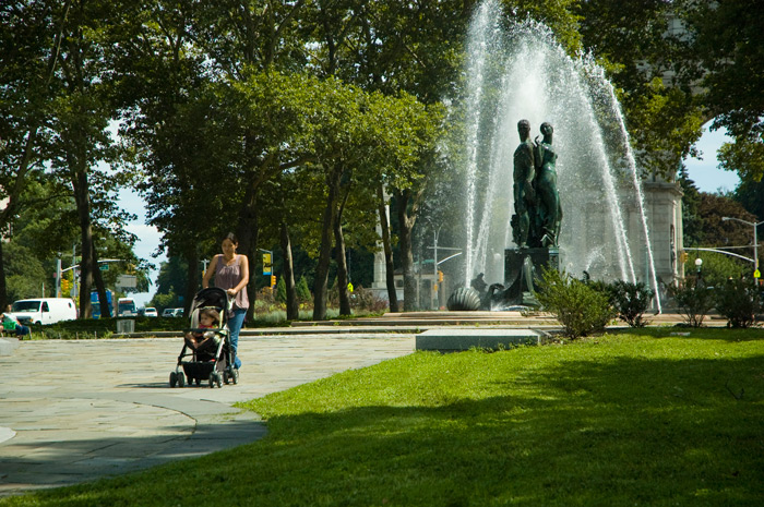 A woman pushes a baby stroller through a plaza with a statue-filled water fountain.