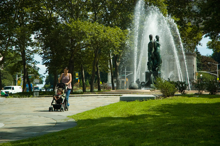 A woman pushes a baby stroller through a plaza with a statue-filled water fountain.