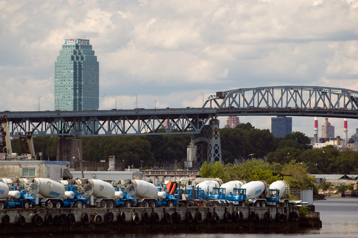 A parking lot full of cement trucks sits in front of an overpass and a lone sky scraper.