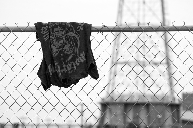A black t-shirt hangs over the top of a chain-link fence, with the tower of an old amusement park ride in the distance.