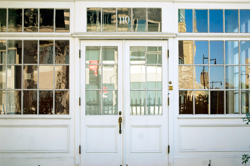 An outer vestibule like a sun room, with French doors and many window panes, reflects the surrounding buildings and the blue sky.