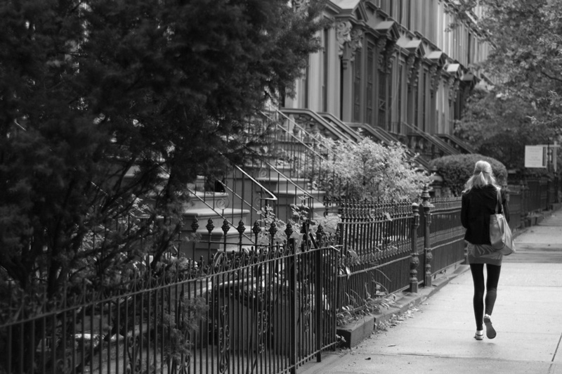 A woman walking down the sidewalk on a street filled with brownstones.