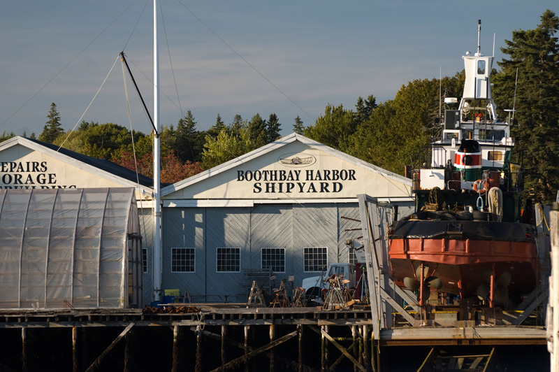 A red wooden tugboat is suspended in dry dock at a small shipyard.