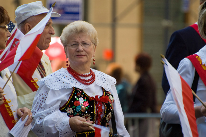 An older woman dressed in traditional folk costume waves a Polish flag.