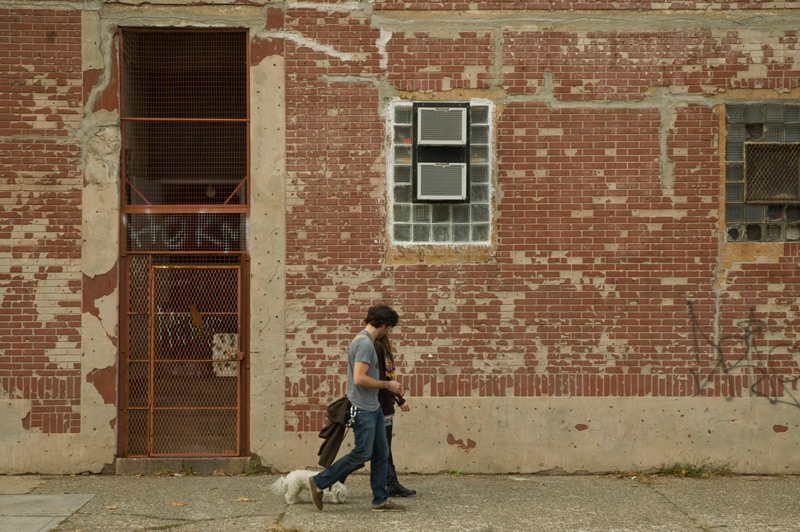 A couple walks their dog past an old brick building.