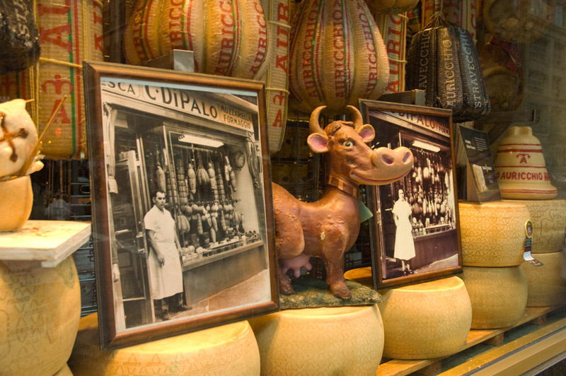 A delicatessen's window display consists of cheese wheels, a cartoonish cow, and founders' pictures.