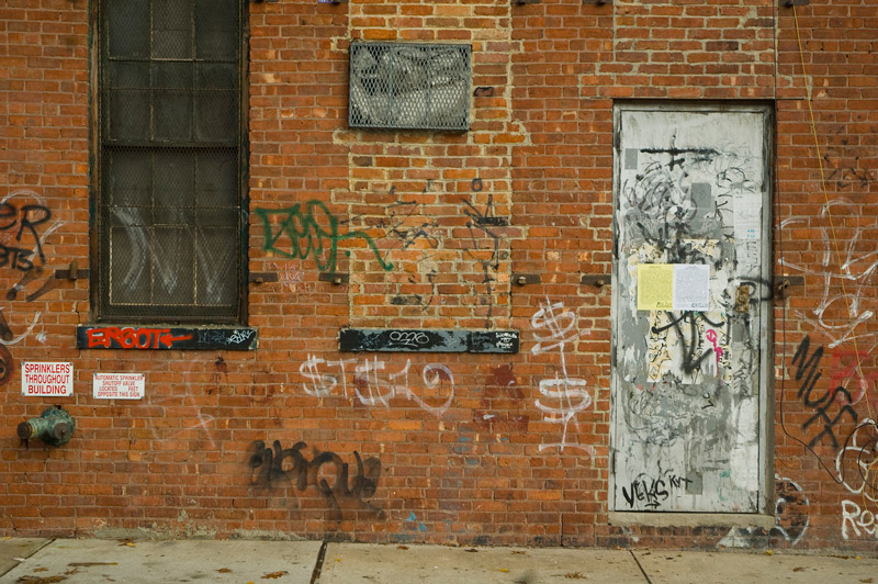 A brick wall and a door are covered with graffiti rags, the door also has two sheets of paper posted on it.
