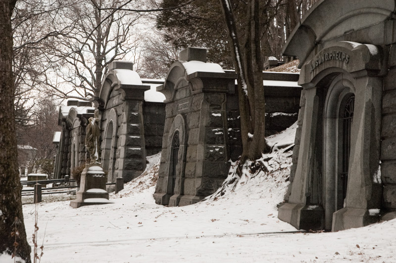 A row of mausoleums, with snow and bare trees.