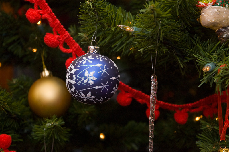 A frosted blue ornament hangs in front of red garland on a Christmas tree.