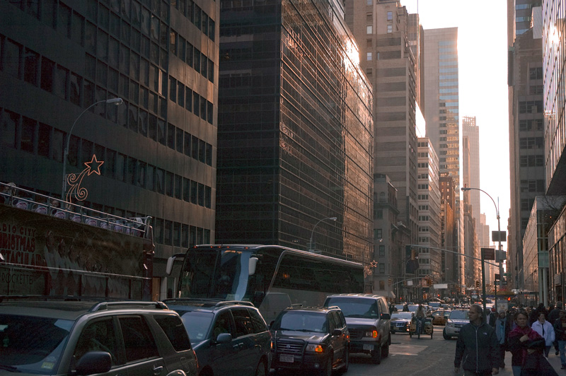 Cars and buses line up to stop lights between a canyon of skyscrapers.