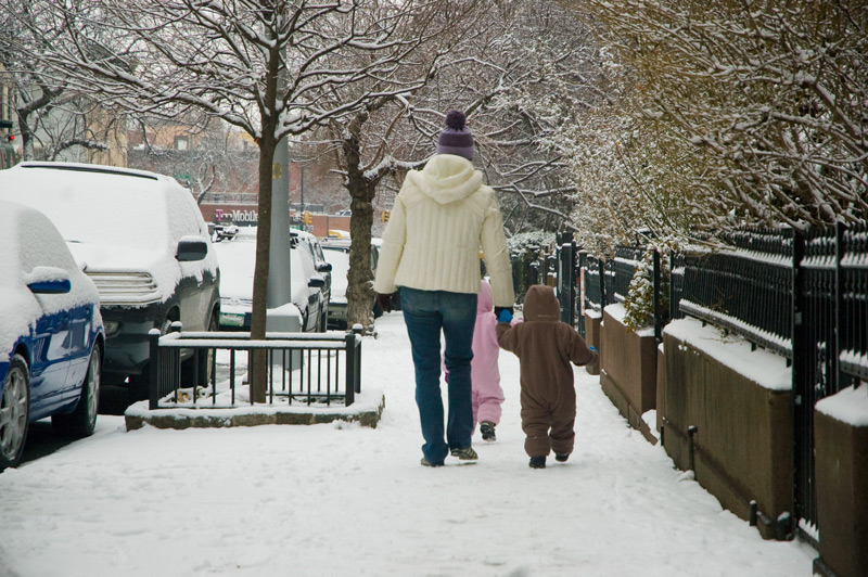 A mother holds her toddlers' hands as they walk down a sidewalk, with snow on fences, cars, and bushes.