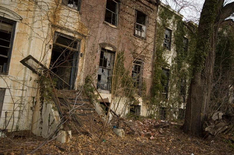 Abandoned row houses with broken windows, surrounded by weeds.