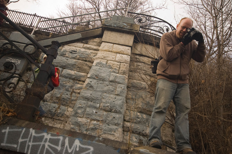 A photographer squints through his viewfinder while standing on a ledge below stonework.