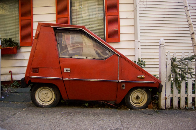 A small, angular, red electric car sits in front of a white house with red trim.