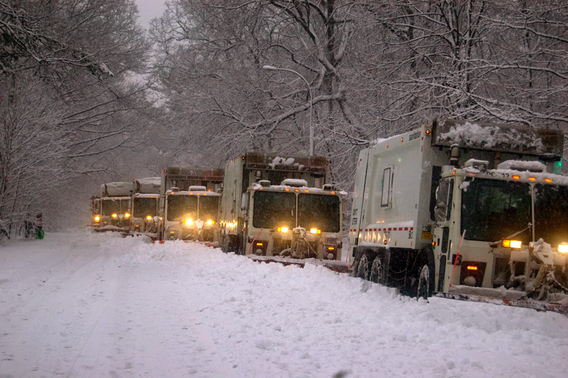 A half dozen garbage trucks with snow plows on their front are lined up to clear a wintry road.