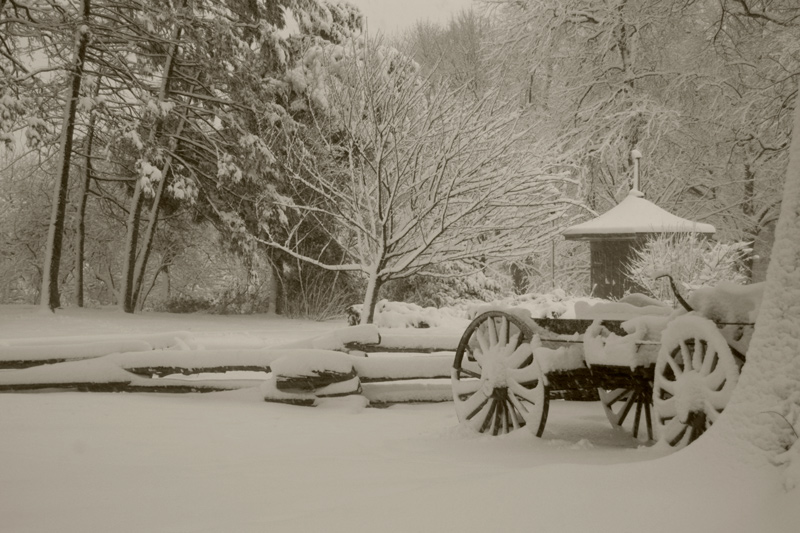 Outlines of a wagon peek through the snow by a log fence.
