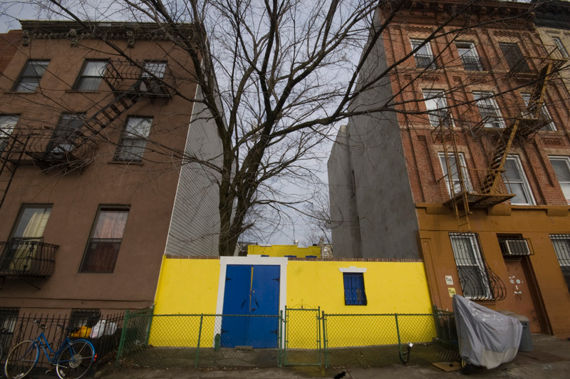 A blue and yellow wall provides privacy for a yellow, setback house.