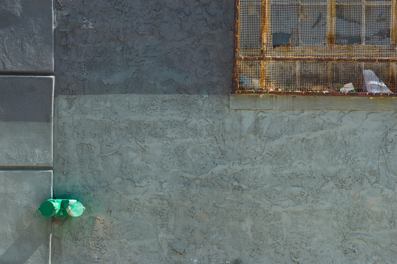 A blue-grey stucco wall, broken by water pipes and a rusted window grate.