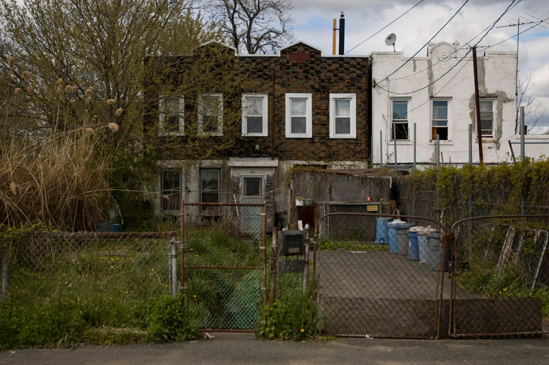 Behind chain-link fence, an overgrown lawm, a driveway, and a modest house.