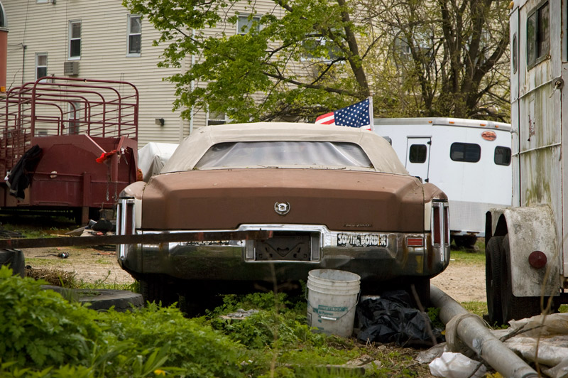 A rusting old car, sitting by horse stables.