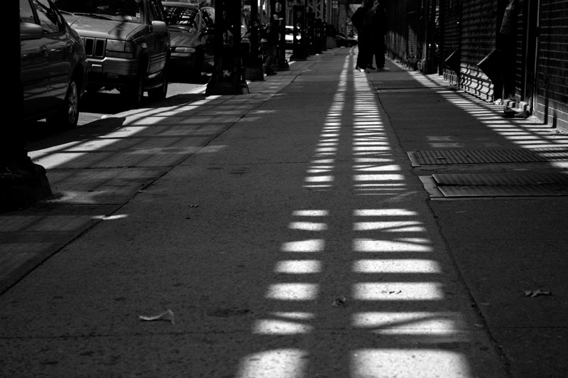 Shadows from an elevated train track dominate a sidewalk.