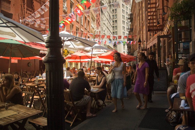 People eat at wooden tables wedged between tall buildings.