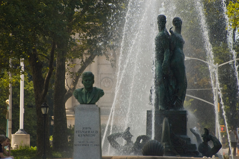 A bust of JFK, in front of a water fountain with two tall figures.