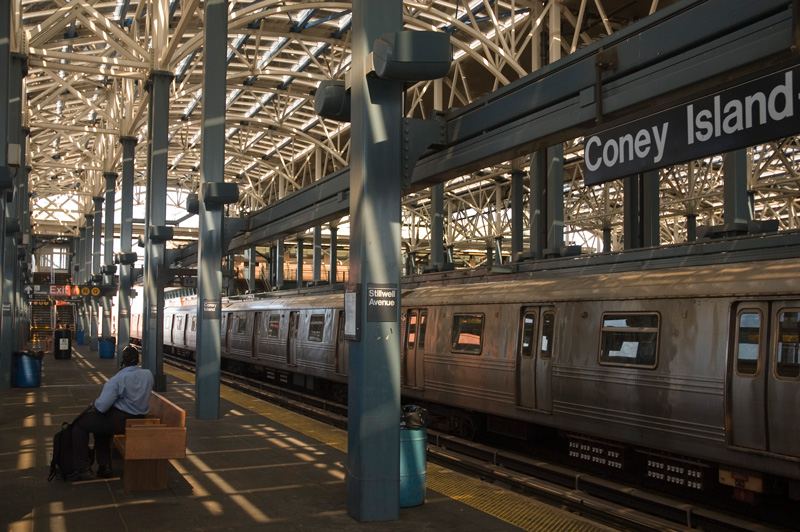 A man on a bench on a subway platform with a networked open-air ceiling.