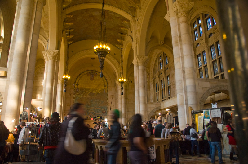 A room with high ceilings, columns, chandeliers, mosaics, and people at a flea market.