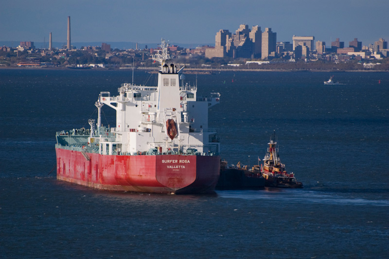 A tug boat guides a ship through the harbor, Brooklyn in the background.