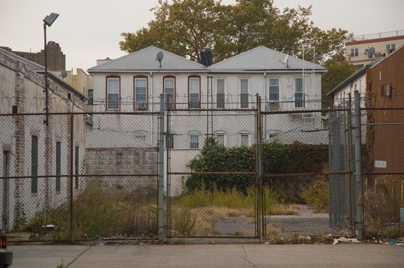 A weed-overgrown lot, behind chain link fence.