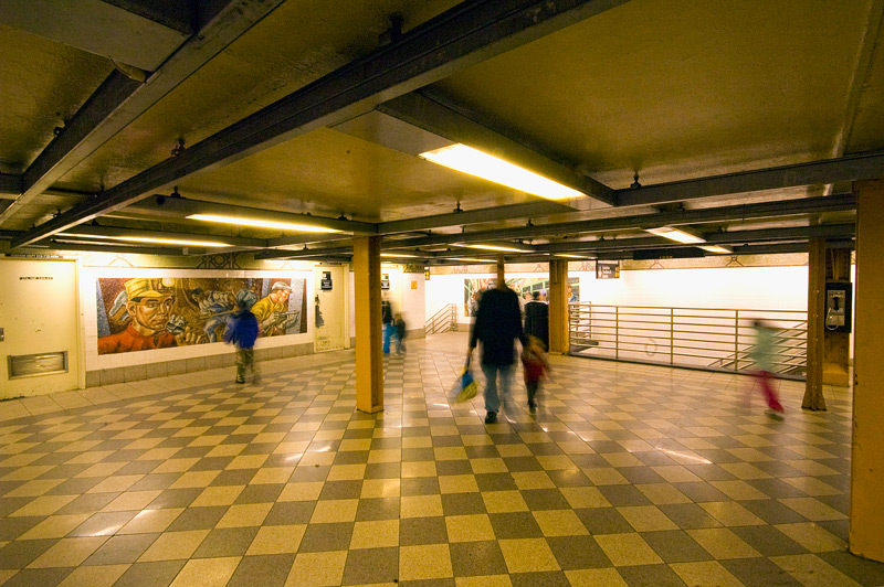 A subway mezzanine, with checkered floor tiles, mosaics, and a few people.