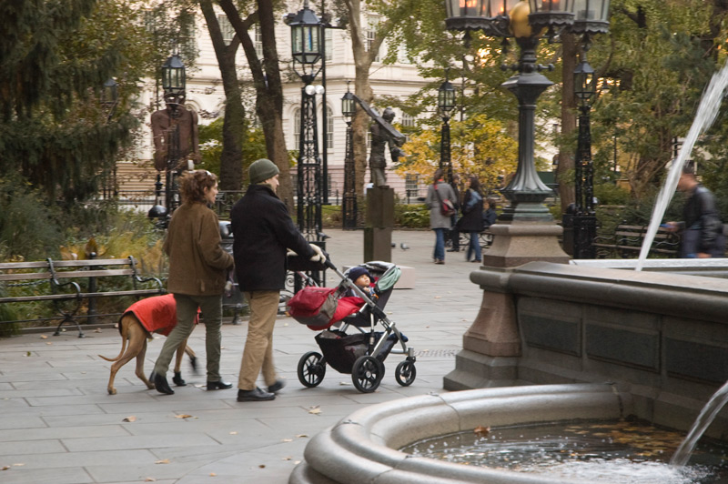 A couple with a baby stroller pass a water fountain, and the child watches the fountain.