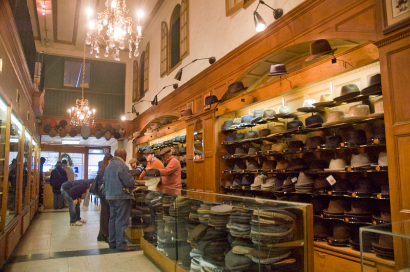 Customers and attendants on opposite sides of a counter, in a room filled with hats.