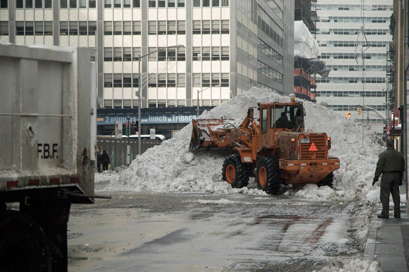 A steam shovel works at a huge pile of snow, between skyscrapers.