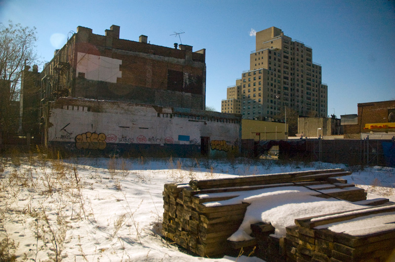 A snow-filled construction lot, with tall buildings beyond it.