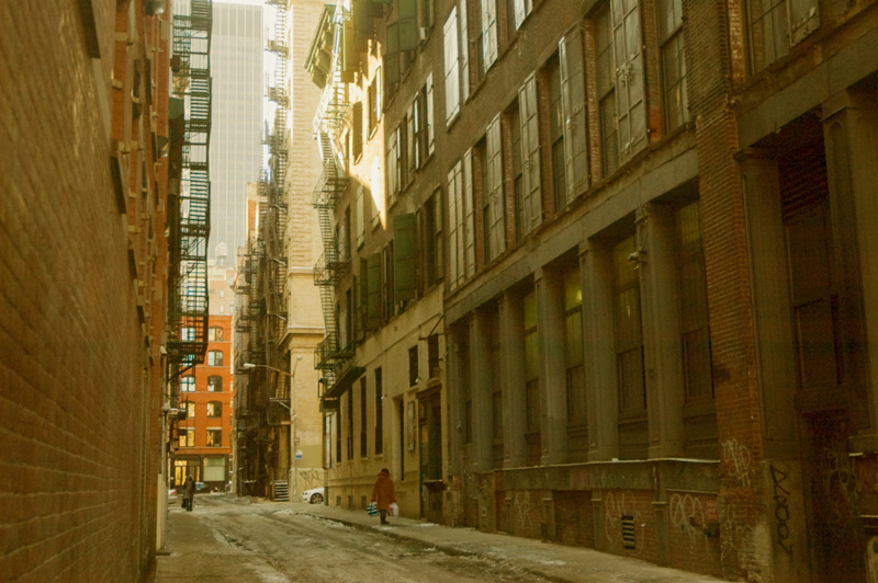 A woman walks down a thin alley with many fire escapes.