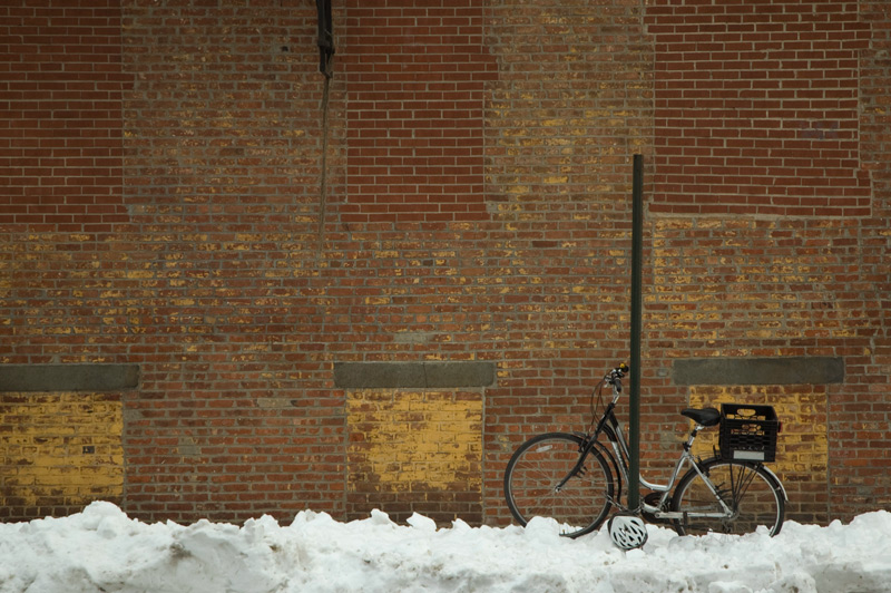 Snow on the sidewalk, a brick wall, and a bike locked to a post.