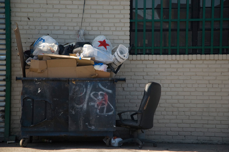 An old office chair sitting next to an over-stuffed dumpster.