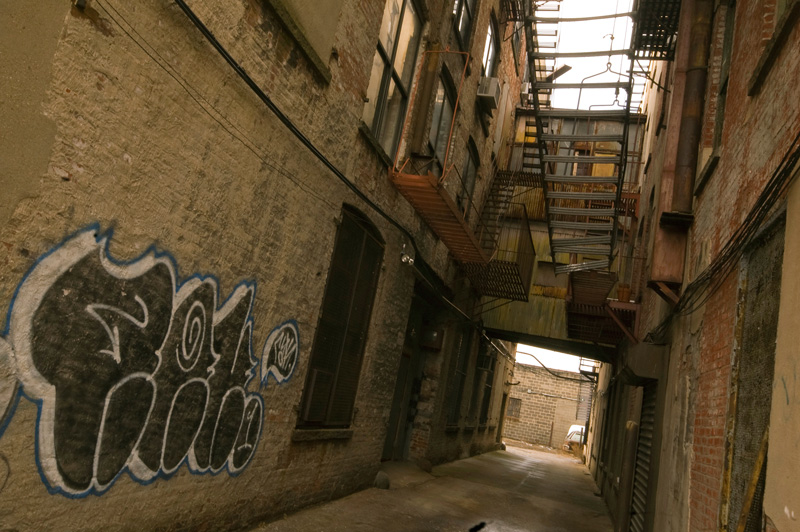 A tunnel-like alley, with overhanging fire escapes